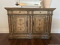 Front view of antique style buffet cabinet showing two drawers, two cabinet doors with floral painted panels, fluted columns and decorative metal hardware.