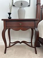 Front view of walnut antique style end table showing single drawer and curved legs with cross stretchers, placed on carpet with lamp and decor on top.