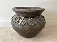 Front view of the grey clay jardiniere planter showing metallic floral and swirl designs around the rim, on carpeted floor against white wall.