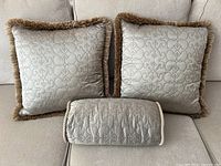 Set of two square taupe decorative pillows with fringe and one taupe bolster pillow on a light sofa.