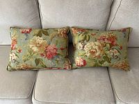 Pair of floral chintz sage green decorative pillows shown on a light gray couch, front view showing full surface detail with floral pattern including cream roses, pink peonies, blue lilacs and green leaves.