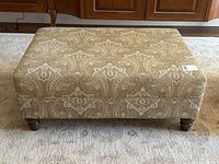 Front view of large rectangular ottoman with beige paisley upholstery and turned wood legs.