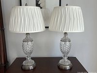 Pair of Dale Tiffany crystal lamps shown on tabletop with white pleated lamp shades and brushed metal bases.
