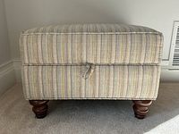 Front view of striped fabric upholstered ottoman with dark turned wooden legs and storage lid with fabric loop pull