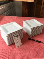 Two stacks of white square tapas plates on a pink tablecloth with a pen for scale and a MaxSold tag labeled '700' and 'Tapas Plates'.