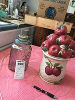 Photo showing the ceramic apple-themed cookie jar with apples on lid and woven detail next to the carnival glass cookie jar on a pink crocheted tablecloth.