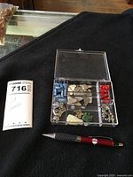 Photo of a clear plastic box with compartments showing cat beads and pendants in various colors and styles with a pen for scale.