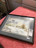 Framed watercolor painting showing a snow-covered winter landscape with bare trees and buildings in the background, double matted under glass.