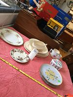 Overview of the floral patterned dishes, plates, teacup, sugar bowl, and decorative tin on a pink cloth with measuring tape visible for scale.