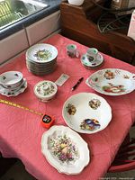 Photo of fruit motif dishes including plates, saucers, cups, bowls, ramekin, and tray on pink cloth on table with tape measure and pen for size reference.
