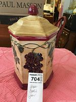 Full shot of hexagonal grape motif cookie jar by Nonnis with lid and maroon trim on pink cloth.