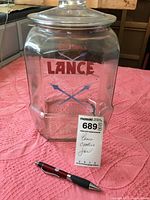 Full view of the Lance cookie jar on a pink textured fabric. Jar is octagonal, clear glass with the Lance logo and crossed arrows printed on the front.