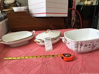 Three large porcelain casseroles with two handles each displayed on a pink textured tablecloth, a measuring tape showing size in front.