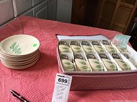 Photo showing the set of 12 cups with two handles and 12 saucers stacked and boxed, laid on a pink fabric surface.