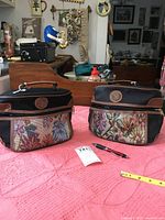 Two identical black luggage totes with brown trims and a multicolor floral brocade pattern on the bottom portion, placed side by side on a pink quilted surface.