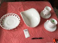 Photo showing the three milk glass items spread out on a textured pink cloth: a round pierced edge bowl, a rectangular wavy fluted bowl, and two coffee cups each on a saucer with a grapevine design.