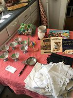 Overview of assorted miscellaneous items on a pink surface including glassware, linens, books, cookie jar, pewter bowls and small toys