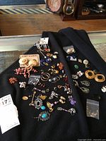 Photo showing the full collection of various pierced earrings spread out on a black fabric surface including hoop, dangle, stud, and clip-on styles with diverse colors and materials.