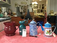 Overview photo of four pitchers on counter including red Longaberger, green pottery, blue and white splatter ceramic, and colorful base metal with rooster design.