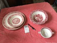 Full view of all red transferware items including plates, bowls, and handled soup cup on a pink fabric surface.