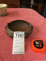 Top-down angled view of a dark toned Roseville Florentine ceramic bowl with handles, placed on pink fabric with a tape measure next to it.