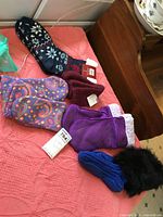 Five pairs of ladies slipper socks displayed on pink fabric, including blue-black knit with snowflakes, two purple fleece pairs, maroon fleece pair, and paisley patterned fleece pair.