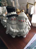 Front and side view of a ceramic cookie jar shaped as two snowmen figures standing together, one with a black top hat and the other with a white winter hat with gold trim. They have scarves and mittened hands and detailed facial features, approximately 12 inches tall and 10 inches wide.