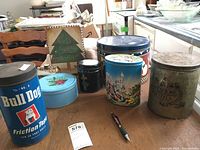 Photo of six assorted collectible tins on wooden table with chairs and kitchen counter visible.