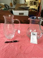 Two clear glass pitchers on a pink cloth surface, one tall with etched bottom, the other wide and plain glass.