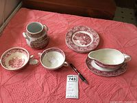 Photo of the entire group of Wedgwood red transferware items including plates, soup bowls, gravy boat with underplate, and small vase on a red textured fabric.
