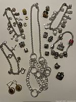 Top-down view of three silver-tone charm bracelets and one silver-tone chain necklace placed on table with assorted individual charms and clips scattered around them.