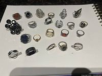 Group view of 21 fashion rings arranged on white background showing variety of designs and stones including black, amber/orange, green, pearl and clear rhinestones.