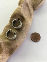 Two small sterling silver hoop earrings displayed on a textured wooden holder next to a penny for size comparison. Earrings have a diameter of about 1.5cm and show tarnish and surface wear.