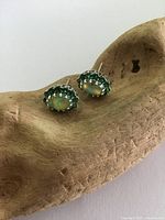 Close-up image showing oval opals surrounded by green rhinestones set in sterling silver, presented on a carved wood background.