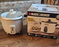 Photo of large Black & Decker rice cooker next to its box on table, shows cooker with white body, black side handles, glass lid, and control switch.