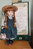 Full view of porcelain doll wearing floral blouse, green pinafore dress, straw hat, black shoes, holding fabric bag, standing on display stand with original box behind.