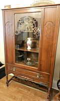 Front view of antique china cabinet showing glass door with decorative carved wood panel inside and contents behind glass.