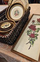 Basket holding four framed needlework pictures with floral designs, viewed from angle