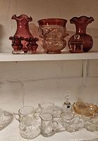 Shelf arrangement of cranberry red vases, bowls and small decorative pieces on the top shelf; assorted clear glassware including pitchers, bowl and decanter on the lower shelf.