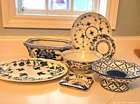 Photo showing 7 assorted blue and white decorative bowls and plates arranged on a white counter in front of a window, displaying various floral and geometric blue patterns on white ceramic.