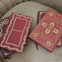 Four classic books stacked on a couch, showing red and burgundy leather covers with gold embossing and decorative patterns.