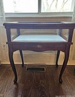 Front view of the wooden display table with glass top and glass sides, placed in front of a window on a wooden floor.