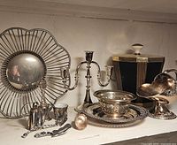 Shelf displaying a round silver plated bread basket, two-arm candelabra, ornate silver plated bowl on tray, black gold container with knob lid, and silver plated sauce boat, plus smaller silver items