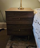Front view of wood grain 2 drawer night table with metal round knobs standing on metal legs beside a bed.
