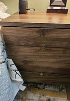 Night table front showing two drawers with wood grain finish and metal knob handles, standing near a bed.