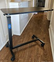 Front angle view of the full adjustable over bed table, showing black metal frame on wheels, and rectangular wood grain surface top.