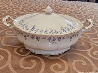 Side view of the covered serving dish showing scalloped edges, gold trim, blue forget-me-not floral pattern, and handles.