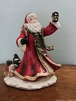 Front and side view of hand-painted Santa Claus figurine in red coat holding a lantern with detailed features and accessories.