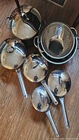 Overview photo showing five stainless steel pots with lids and two colanders stacked together on a wooden floor.