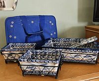 Photo showing three different sized hand painted ceramic casseroles in black metal wire basket holders with blue floral insulated tote bag in background.